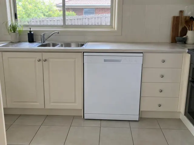 White dishwasher installed under a kitchen benchtop beside a double sink and cream cabinets in Maitland