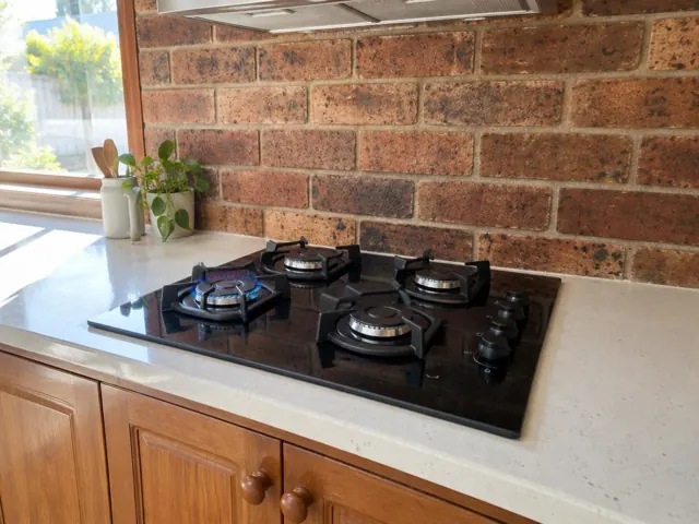 Black gas cooktop set into a light benchtop with brick splashback and timber cabinets in Aberglasslyn