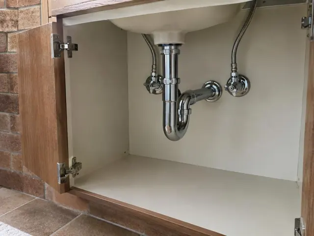 Open under-sink cabinet with neat chrome drain piping and access panel on painted wall in Aberglasslyn