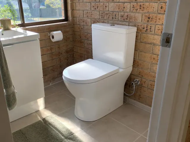 White toilet in a tiled bathroom with brick walls, a window and vanity in Rutherford